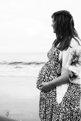 Zwangerschap fotoshoot vrouw kijkt over zee uit met handen op buik in zwartwit